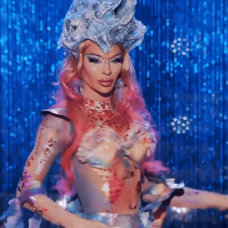 a drag queen with pink hair and body paint is standing on a stage with a blue background .