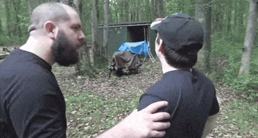 two men are standing in the woods and one of them is wearing a baseball cap