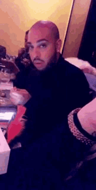 a bald man with a beard is sitting at a table in a room .