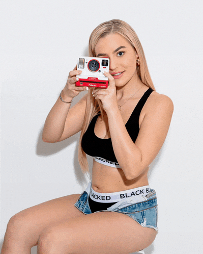 a woman wearing blacked underwear holds a polaroid camera in front of her face