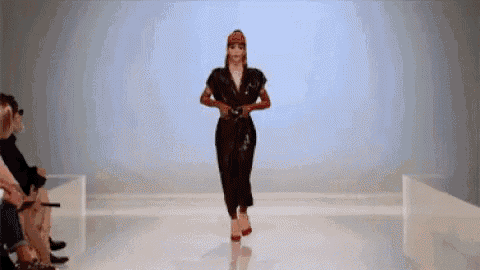 a woman is walking down a runway at a fashion show wearing a black jumpsuit and red heels .