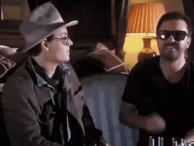 a man wearing a cowboy hat and sunglasses is talking to another man in a room .