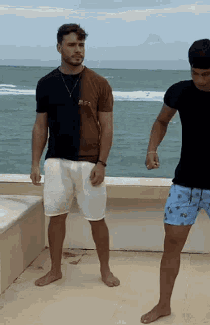 two men standing on a balcony overlooking the ocean with one wearing a black shirt