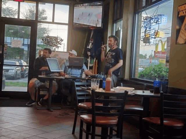 a man is standing in a restaurant talking on a cell phone while sitting at a table with a laptop .