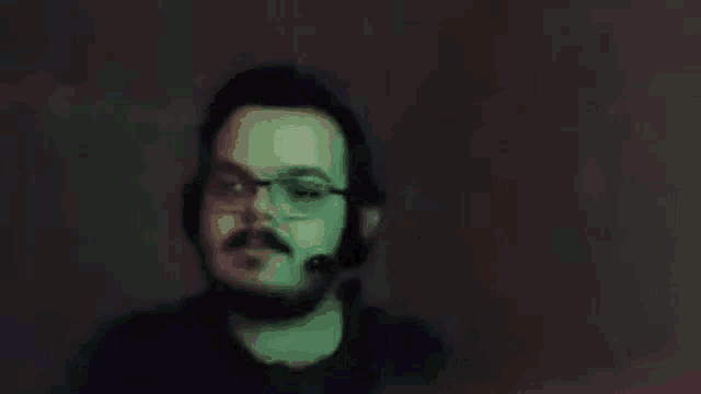 a man with a beard and glasses is wearing headphones and a microphone and pointing at his head .