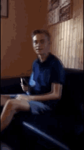 a man in a blue shirt is sitting on a couch looking at his phone