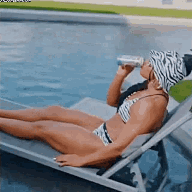 a woman in a bikini is drinking water from a bottle while sitting on a lounge chair next to a pool .