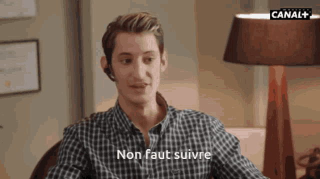 a man in a plaid shirt is sitting in front of a lamp and says non faut suivre