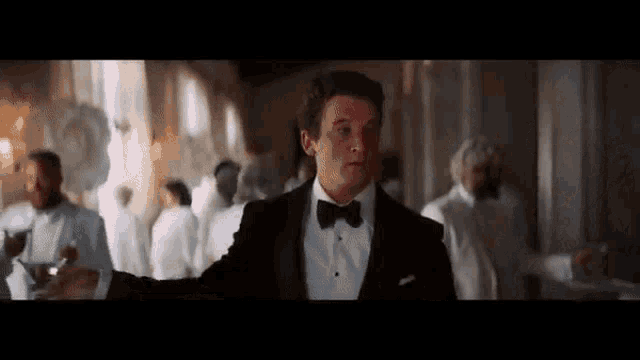 a man in a tuxedo and bow tie is standing in a room with a crowd of people .