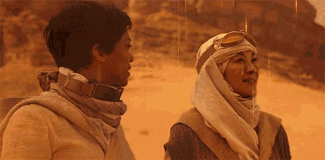 a woman wearing a scarf and goggles stands next to another woman in a desert