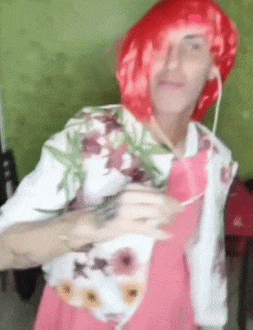 a man wearing a red wig and a pink dress is holding a marijuana plant .