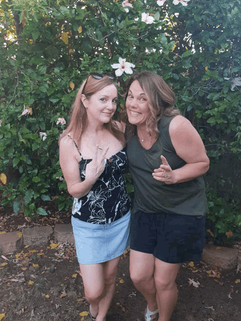 two women pose for a picture in front of a bush with flowers