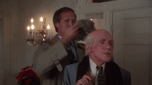 two men are standing next to each other in a room and one of them is touching the other 's hair .