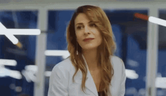a woman in a white coat is standing in front of a window and looking at the camera .