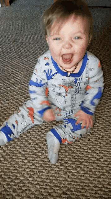 a baby wearing a dinosaur pajamas is sitting on the floor with his mouth open