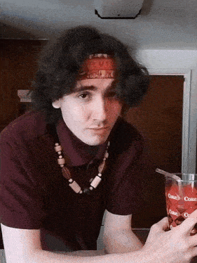 a young man wearing a headband and a necklace holds a glass of coca cola