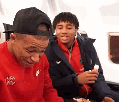 a man wearing a red nike shirt is sitting next to another man wearing a black hat .