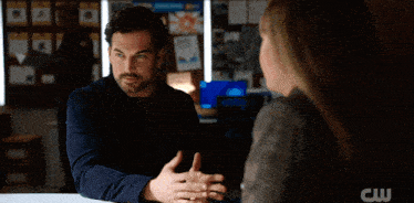 a woman in a plaid jacket is talking to a man with a cw logo on the bottom right
