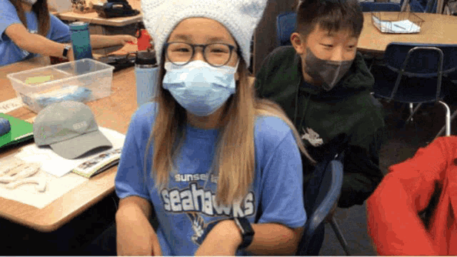 a girl wearing a mask and a seahawks shirt sits at a desk