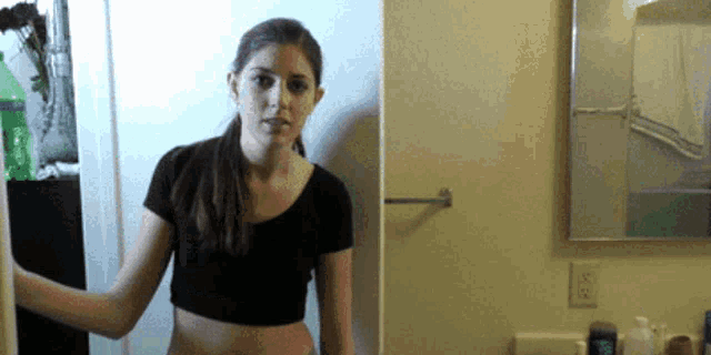 a woman in a black crop top is standing in a bathroom looking at the camera .