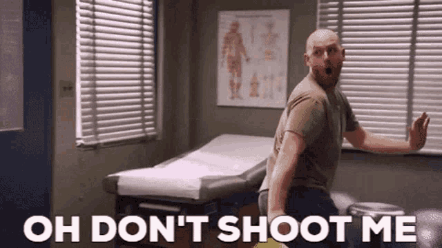 a bald man is standing in a doctor 's office with the words oh don 't shoot me written on the wall .