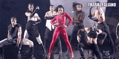 a woman in a red jumpsuit is standing in front of a group of dancers .