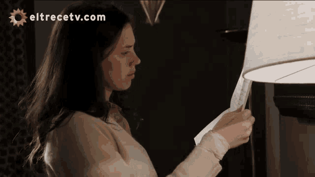 a woman holding a piece of paper in front of a lamp with eltrecetv.com written on the bottom
