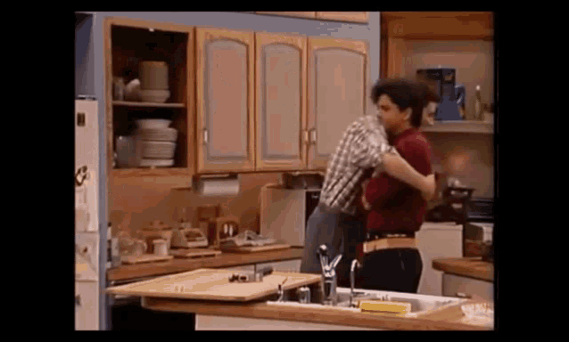 two men are hugging in a kitchen in front of a sink and refrigerator .