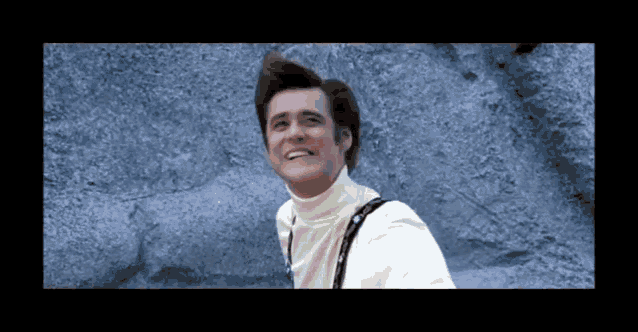 a man in a white turtleneck and suspenders smiles in front of a rock wall