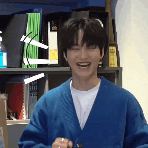 a young man in a blue cardigan is laughing in front of a bookshelf