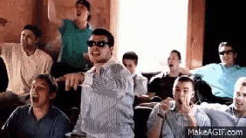 a group of men are sitting in a room watching a movie and one man is pointing at the camera .