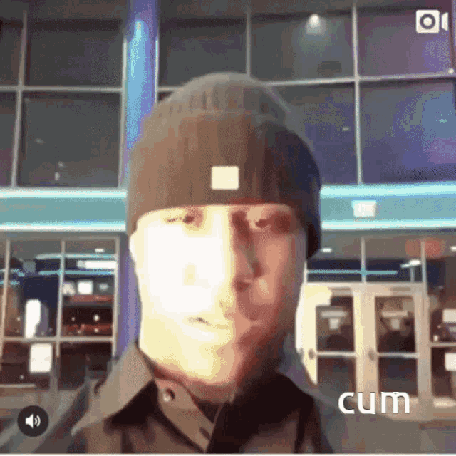 a man wearing a beanie stands in front of a building with the word cum on the bottom right