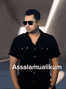 a man wearing sunglasses and a black shirt is standing in front of a sign that says assalamualaikum