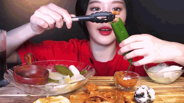 a woman in a red shirt is eating food with tongs and a pickle in her mouth