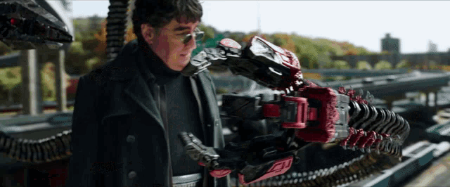 a man in a black coat and glasses is holding a red robotic arm .