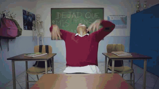 a girl in a red sweater sits at a desk in front of a blackboard that says dejad que mis tans
