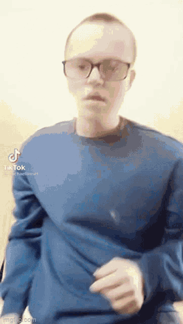 a man wearing glasses and a blue sweater is standing in front of a wall .