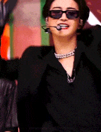 a man wearing sunglasses and a necklace is singing into a microphone .