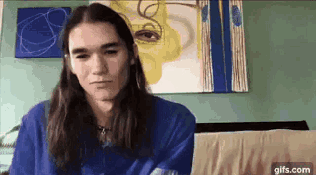 a young man with long hair is sitting on a couch in front of a painting on a wall .