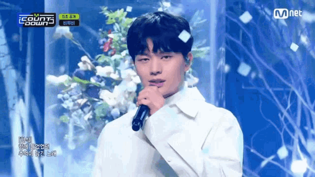 a man singing into a microphone on a stage with flowers behind him