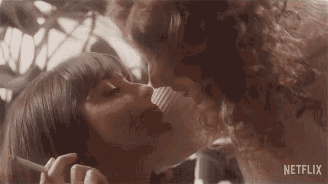 a woman is smoking a cigarette while kissing another woman on the nose .
