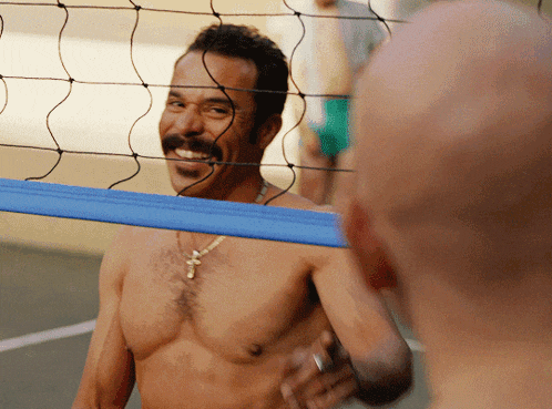a shirtless man with a mustache is smiling at another shirtless man behind a volleyball net