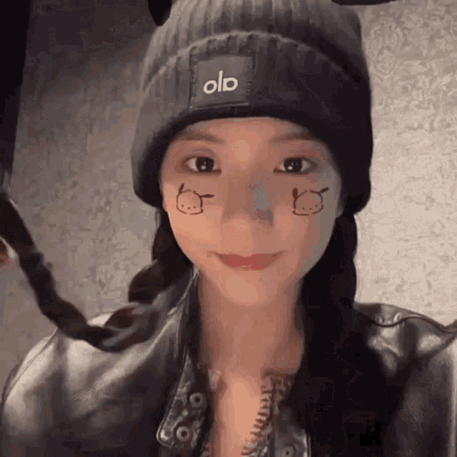 a woman wearing a beanie and a leather jacket has a drawing of a cat on her face .
