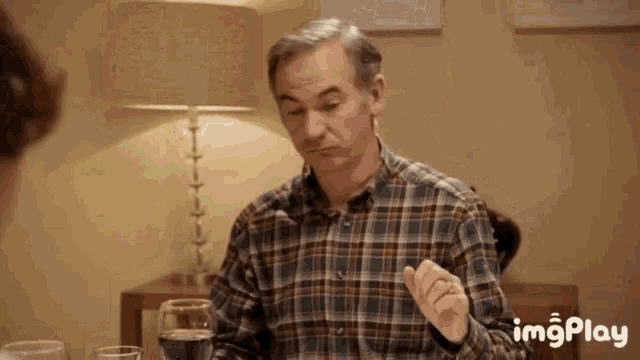 a man in a plaid shirt is sitting at a table with a lamp and a glass of water .