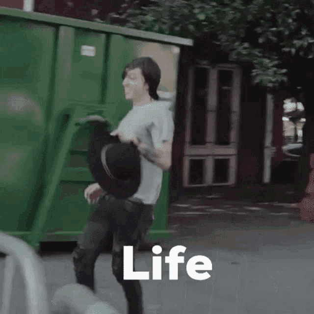 a man is carrying a hat in front of a green dumpster with the word life written on it