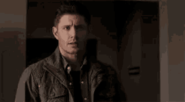 a man in a jacket and black shirt is standing in a dark room .
