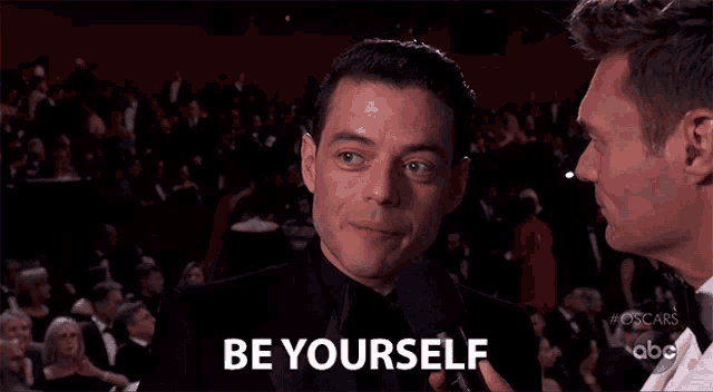 a man in a tuxedo is talking into a microphone and saying be yourself .