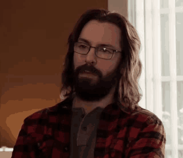 a man with long hair and a beard wearing glasses is sitting in front of a window .