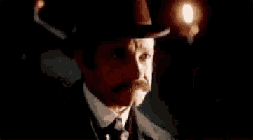 a man in a suit and tie with a mustache is looking at the camera in a dark room .