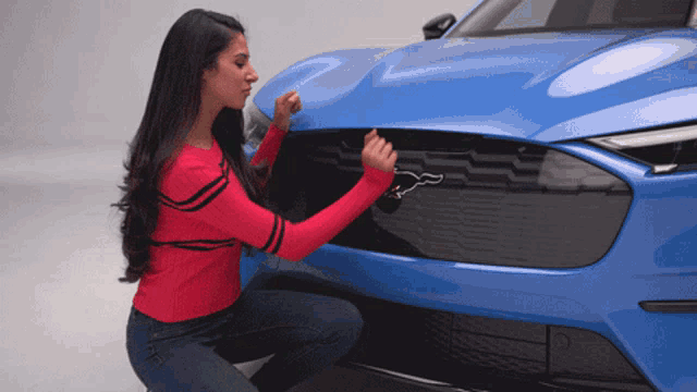 a woman in a red shirt is kneeling down next to a blue mustang